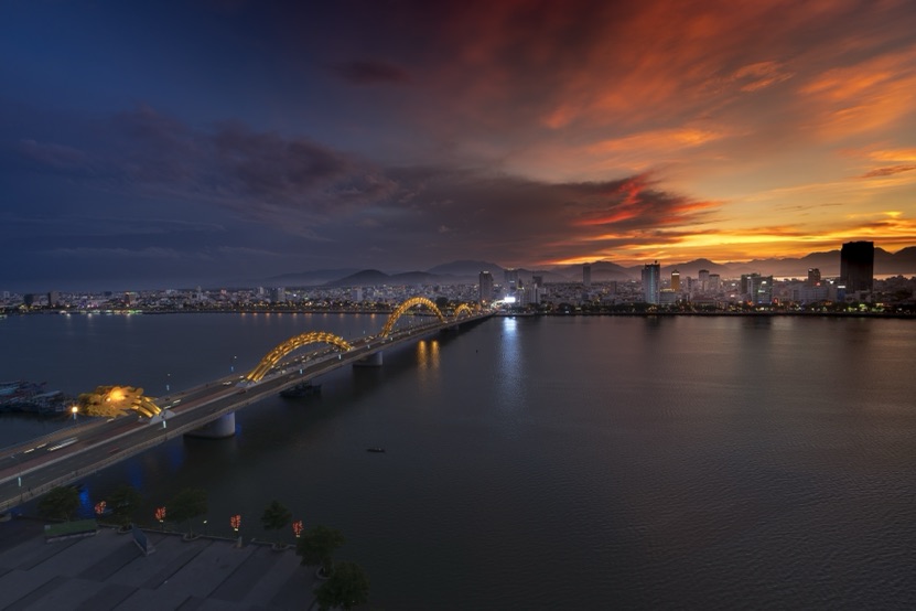 The Dragon Bridge over the Han River in Da Nang at sunset, with the city skyline and the mountains under an orange sky