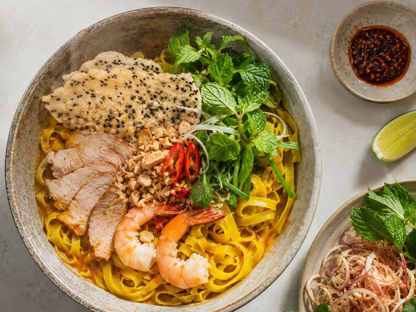 A bowl of mì Quảng with sliced pork, shrimp, roasted peanuts, chilli, mint and a sesame rice cracker, beside a dish of chilli sauce and a plate of herbs and banana flower