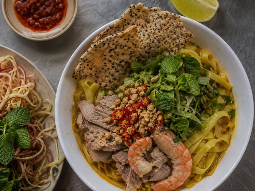 A bowl of mì Quảng with pork, shrimp, peanuts, fresh herbs, chilli and a sesame rice cracker, with a plate of herbs and banana flower beside it