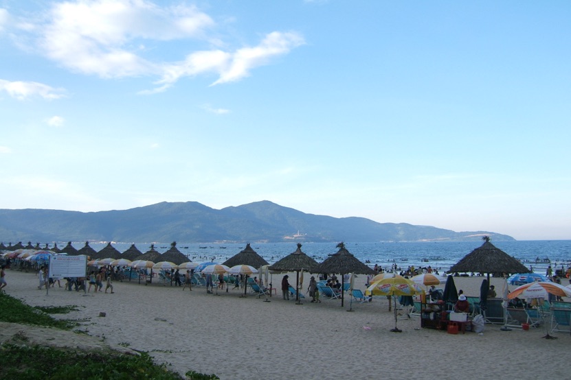 Families under thatched umbrellas on My Khe beach in Da Nang on a sunny afternoon, with the Son Tra peninsula across the bay