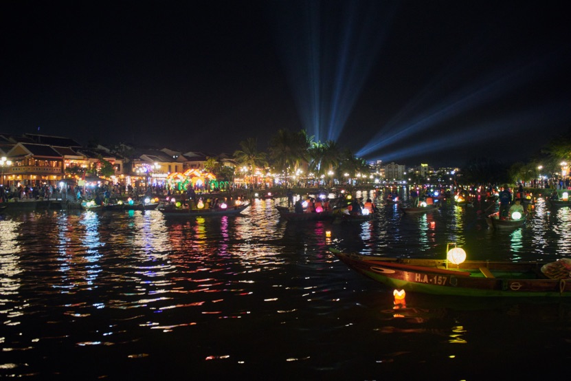 Wooden boats hung with lanterns on the Thu Bon River in Hoi An at night, with the lit old town waterfront behind them