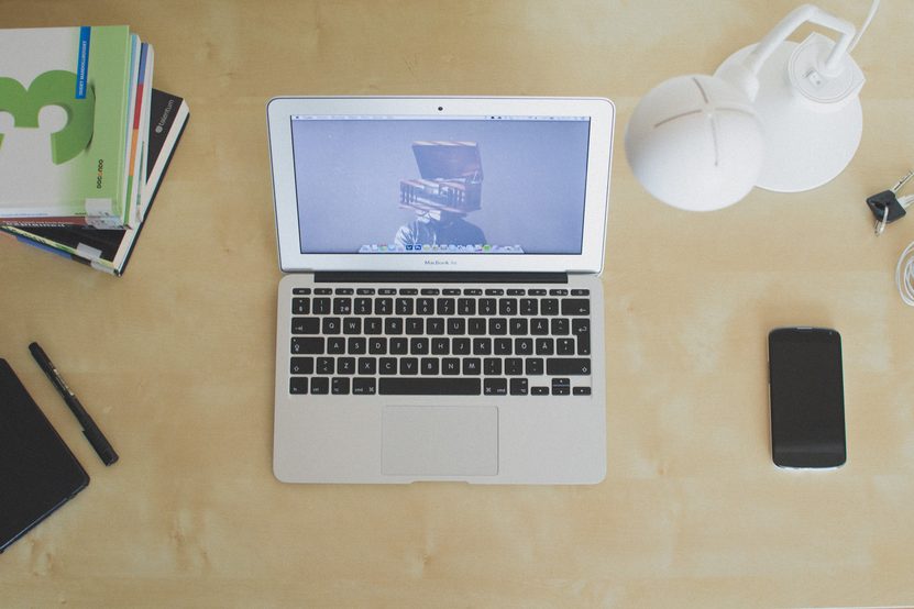 A laptop, notebooks, a desk lamp and a phone laid out on a plain wooden desk